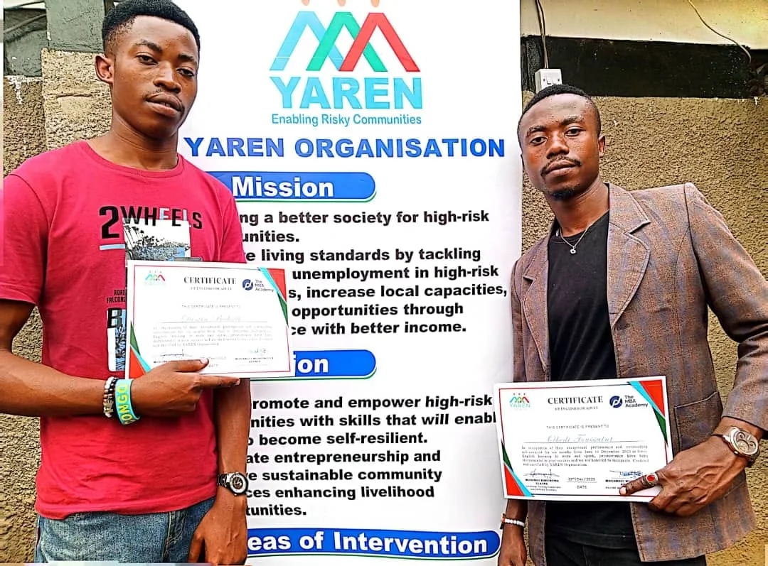 Two YAREN graduates holding their certificates beside the YAREN Organisation banner