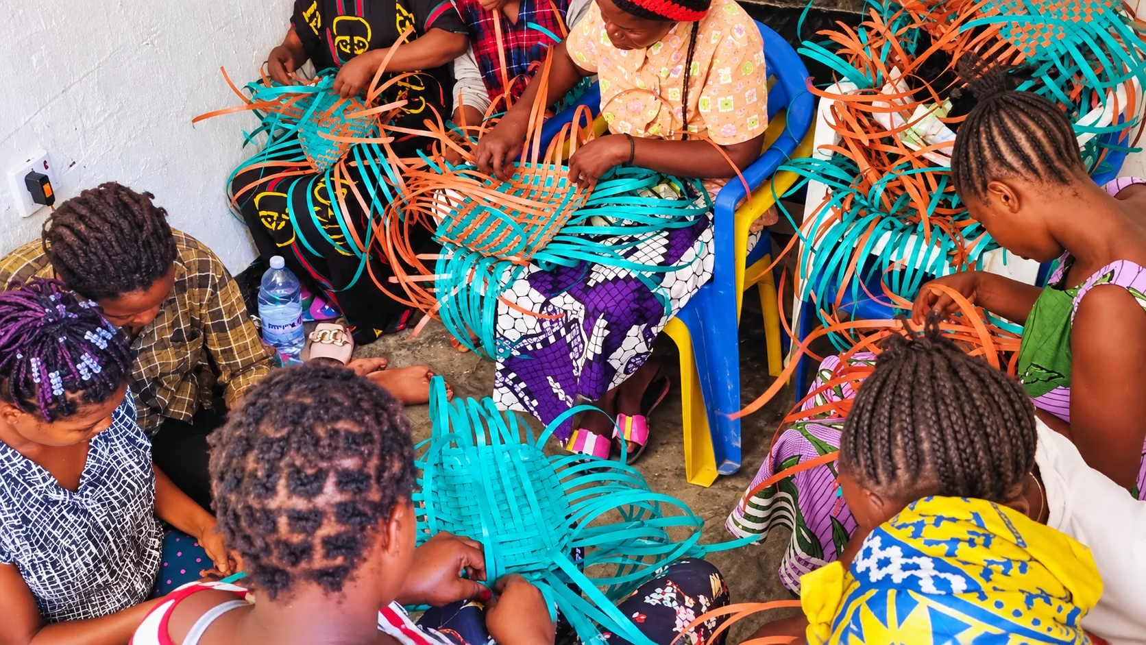 Women sitting together weaving baskets from bright turquoise and orange plastic strips